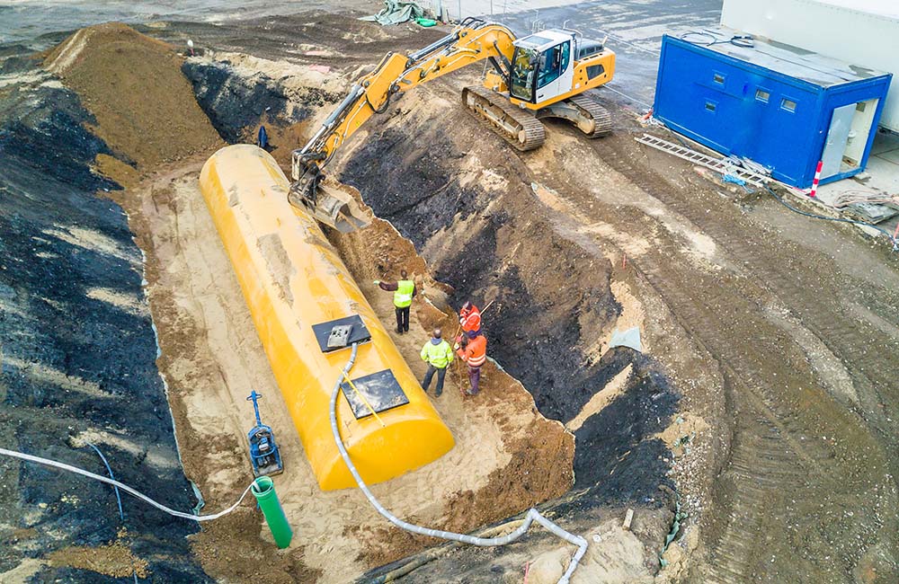 Work crew watching excavator bury a large industrial fuel tank in the ground.