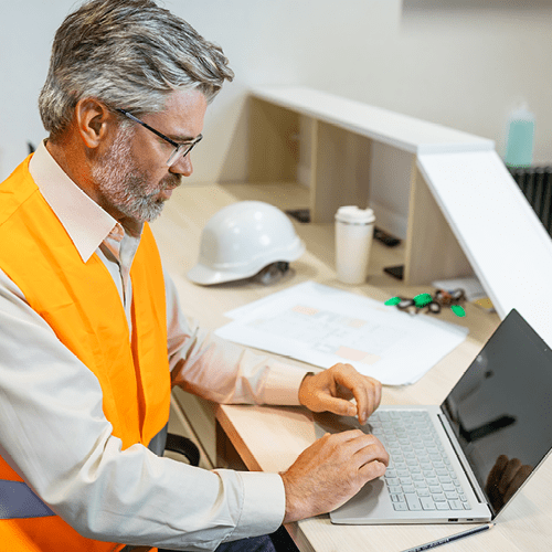 A male engineer in a safety vest working on a laptop.